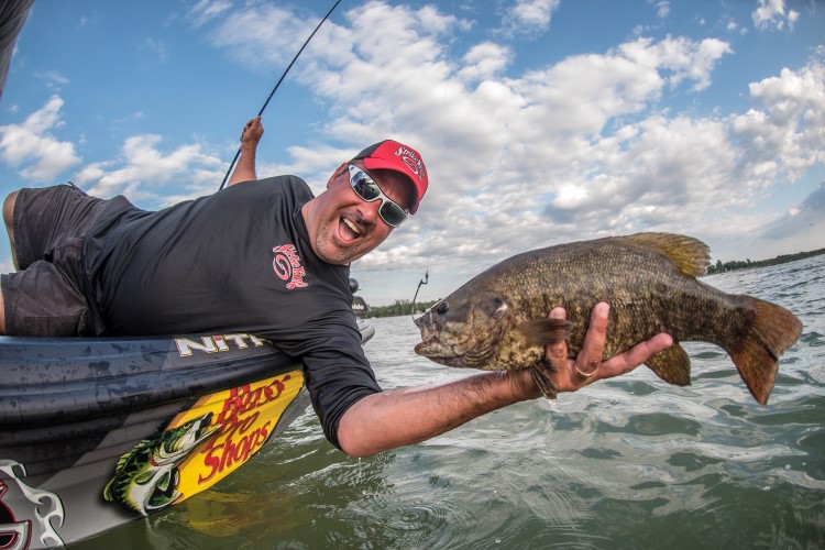 Mark Zona leaning over the side of his boat and showing a smallmouth bass to the camera